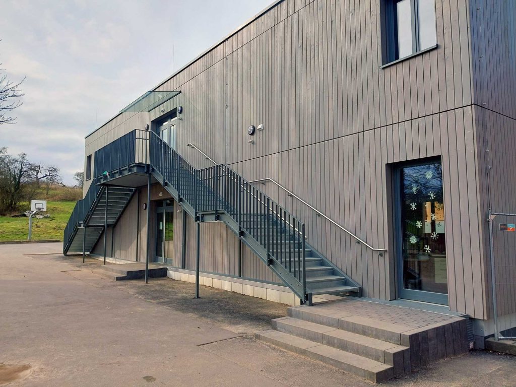 Stahltreppe mit Gitterroststufen, mit Austrittspodest und Zwischenpodesten. An der Außenseite mit einem Flachstahlstabgeländer. Beidseitig mit Edelstahlhandläufen. Alle Stahlkonstruktionsteile feuerverzinkt und farbig pulverbeschichtet.