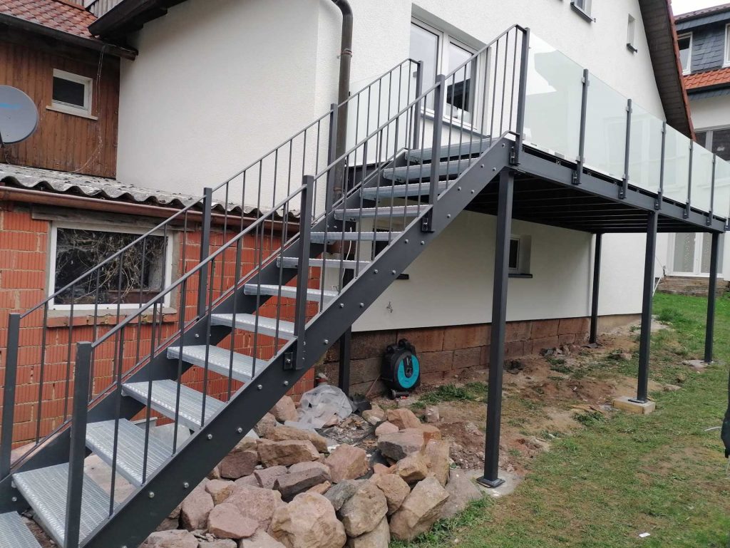 Stahlbalkon mit angebauter Treppe mit Gitterroststufen. Die Gitterroststufen sind verzinkt der restliche Stahl verzinkt und beschichtet. Balkonbelag aus WPC-Dielen. Treppengeländer als Stabgeländer farbig pulverbeschichtet. Balkongeländer aus beschichteten Quadratrohrpfosten mit Milchglasfüllung.