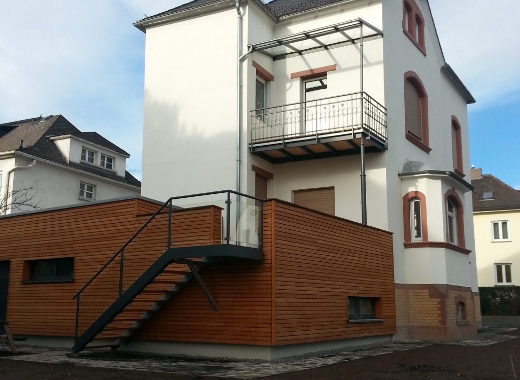 Stahlbalkon auf einer Rundrohrstütze mit Teleskopoptik, Geländer als Stabgeländer mit Schmiedeeisernen Zierelementen, Bodenbelag mit dichtem Flachdachaufbau und Holzdielen