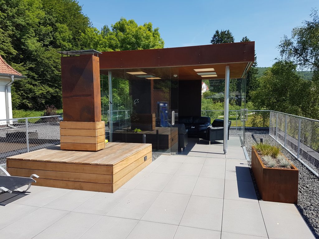 Pavillon mit Cortenstahldach, mit schiebbaren Glasseitentüren. Sitzgelegenheit als Aluminiumgestell mit Holzbrettern beschlagen.