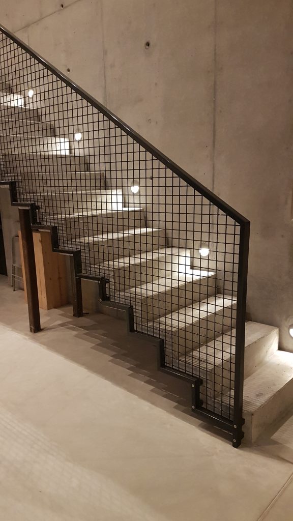 Innentreppengeländer bestehend aus einem Rahmen aus Rechteckrohren mit einer Füllung aus Rundstahlgitter. Aus Baustahl pulverbeschichtet