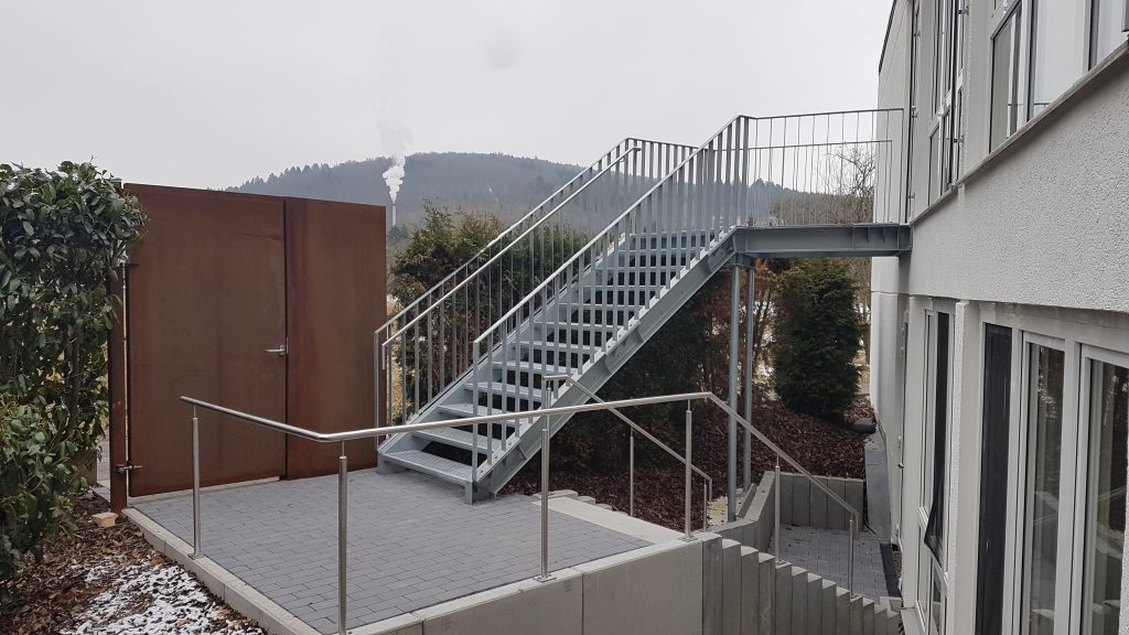 Fluchttreppe aus Baustahl mit Austrittspodest mit Gitterroststufen und Flachstahlgeländer mit nach innen versetzten Edelstahlhandläufen. Cortenstahltüre, Edelstahl Handläufe im Treppenverlauf
