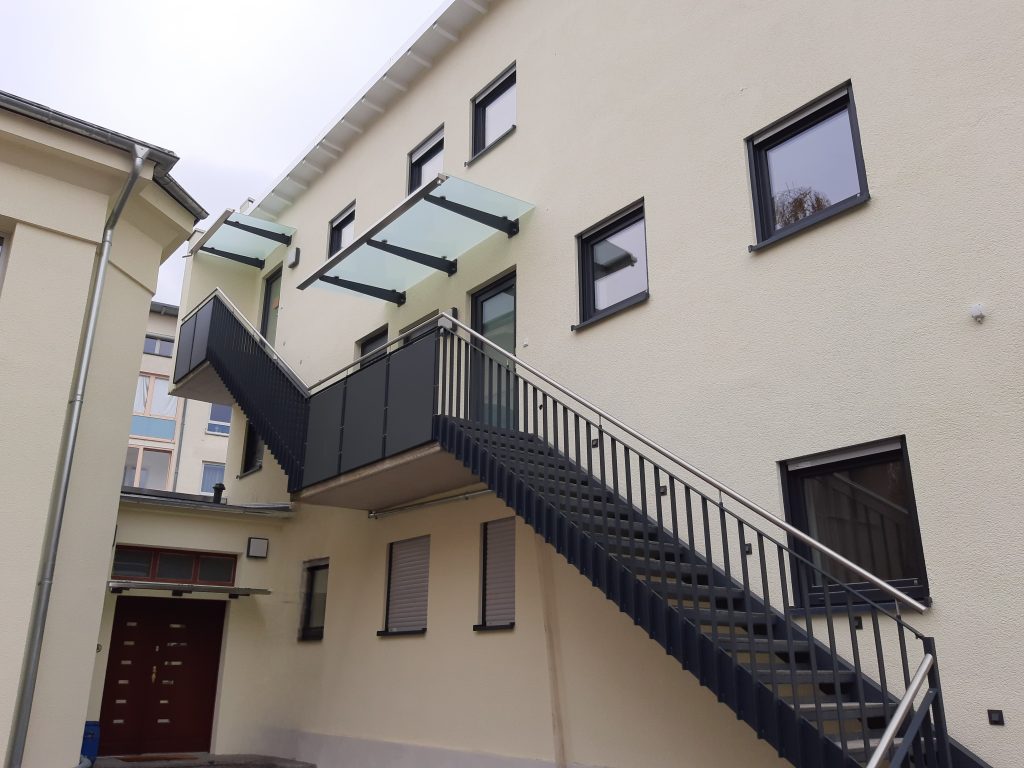 Zweiläufige Außentreppe vom EG bis zum Zweiten OG mit einbindung von Stahlbeton Zwischenpodesten. Mit Stabstahlgeländer mit vertikalen Stäben, im Podestbereich jeweils mit Sichtschutzblechen. Mit aufgesetztem Edelstahlhandlauf und Im Podestbereich jeweils mit Kragarmvordächern mit matten Glasscheiben.
