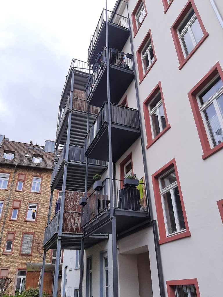 Stahlbalkon 4-geschossig mit Flachstabgeländern und Bodenbelag aus Balkotec-Platten. Stützen aus Quadratrohren und Breitflanschträgern. Stahlteile verzinkt und Pulverbeschichtet im Farbton Anthrazit "Eisenglimmer".