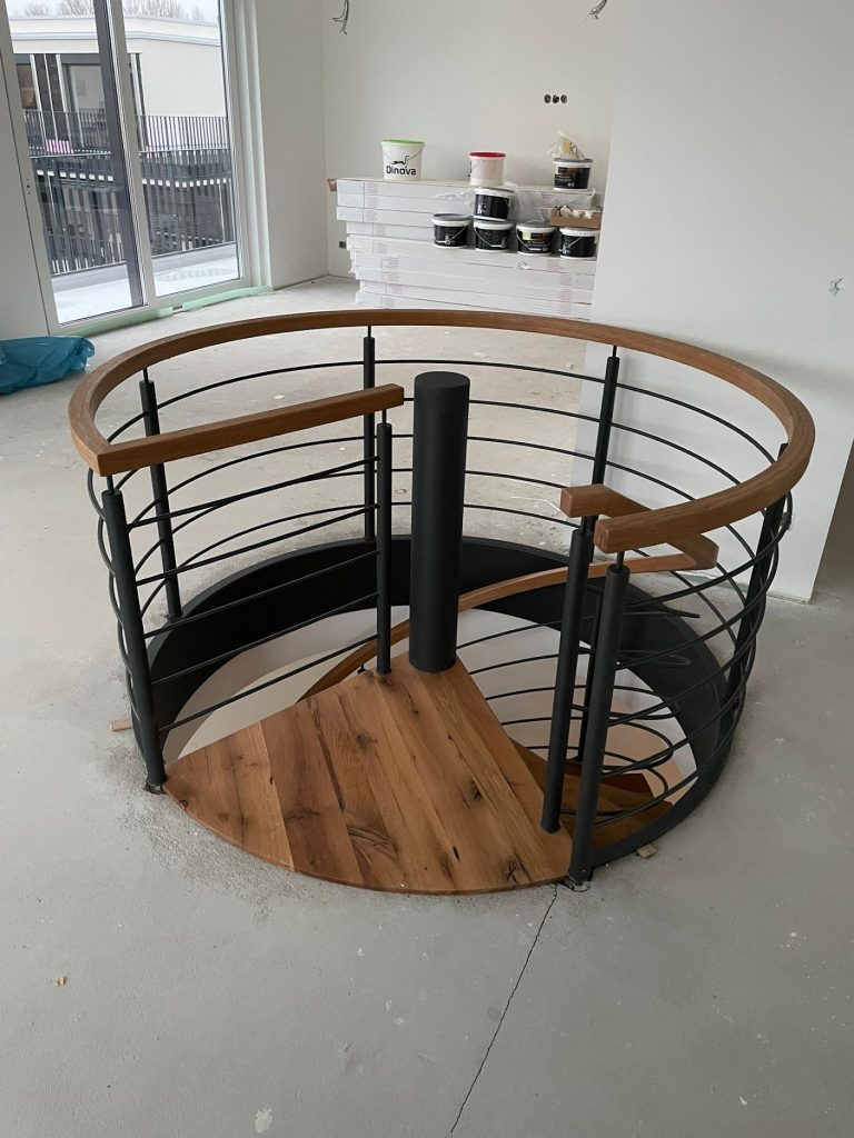 Runde Deckenstirnverkleidung für eine Spndeltreppe mit Geländer aus Rundrohrpfosten und horizontalen Gurten aus Rundstab. mit Holzhandlauf in Stufenart. Alle Stahlteile sind Schwarz-Matt pulverbeschichtet.