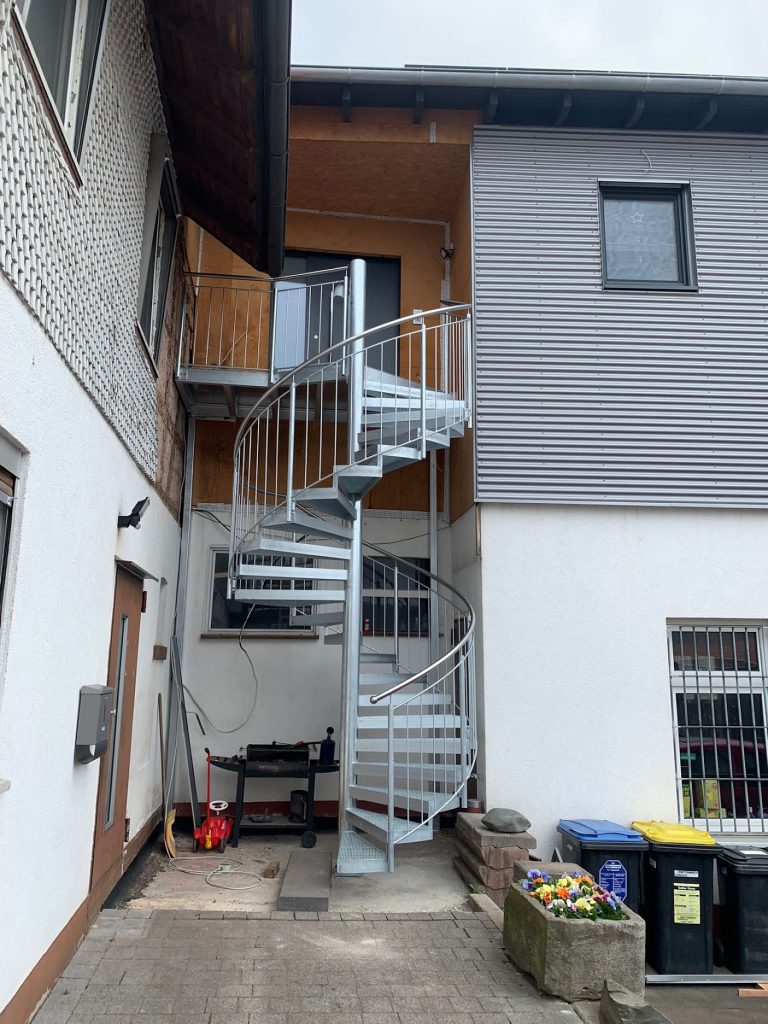 Stahlspindeltreppe für Wohnhausanbau aus einer Spindelsäule mit angeschweißten keilförmigen Spindelstufen und einem Austrittspodest. Geländer aus Rundrohren und Rundstäben. Oberfläche aller Treppenteile feuerverzinkt, Handlauf in Edelstahl Rostfrei.