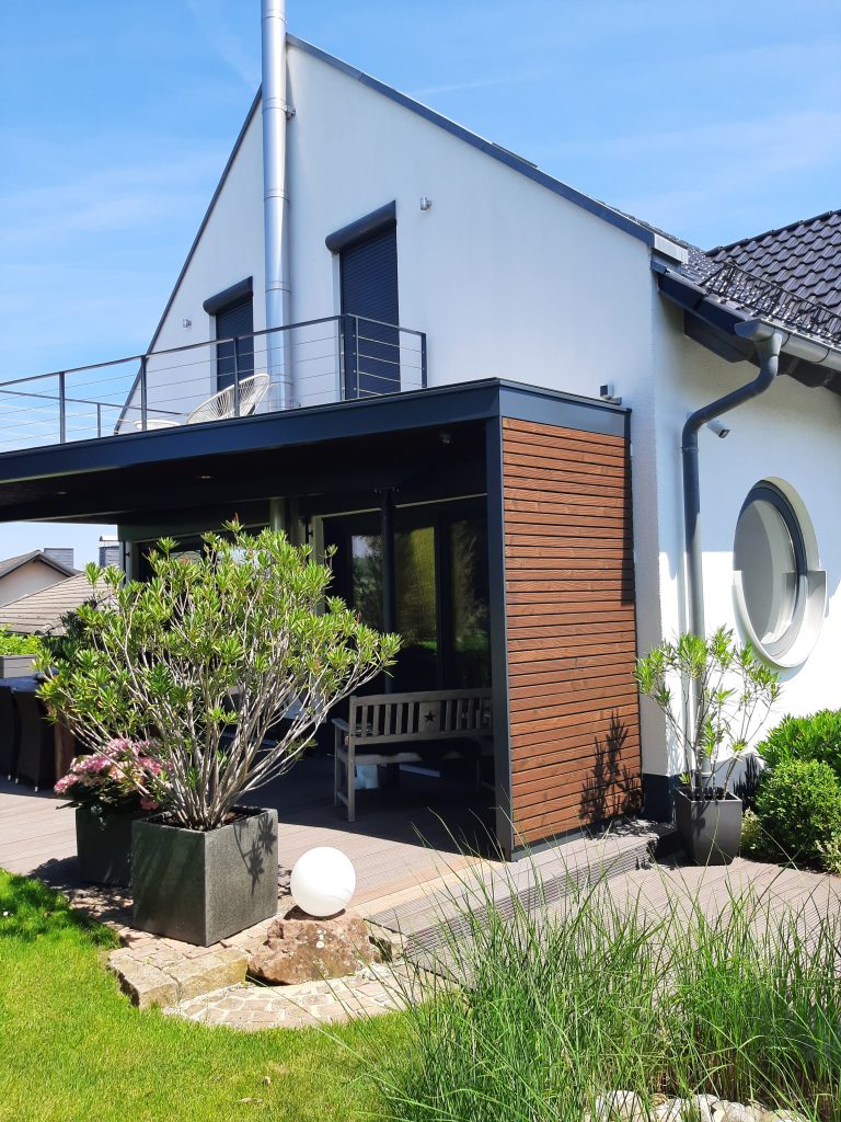 Terrassenüberdachung und Stahlbalkon mit einer, mit Holz beplankten, Seitenwand. Mit einem Geländer aus rechteckigen Pfosten, rechteckigem Handlauf und horizontal gespannten Edelstahl Drahtseilen. Alle Stahlteile sind anthrazit pulverbeschichtet.