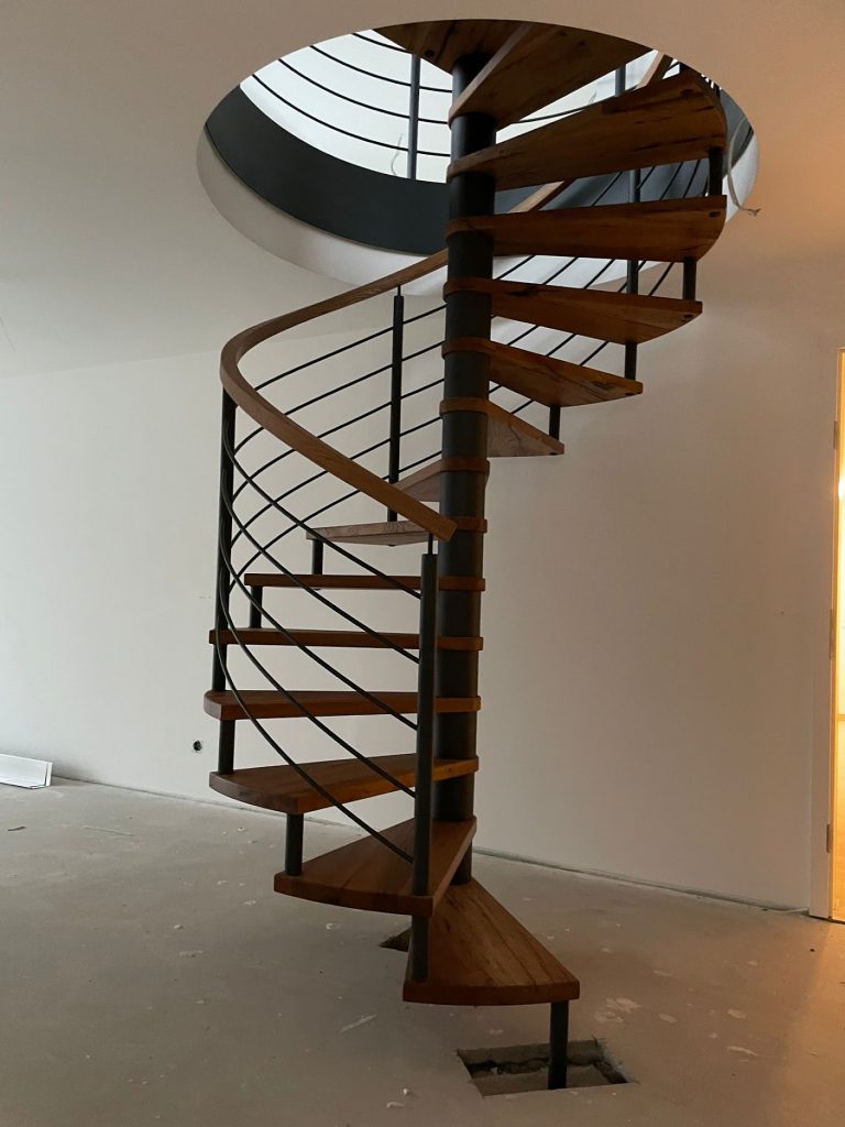 Spindeltreppe aus einer Rohrsäule mit Holzstufen und steigendem Geländer in gewendelter Form mit einem Holzhandlauf und dazu parallel laufenden Gurtstäben. Alle Stahlteile sind Schwarz-Matt lackiert.