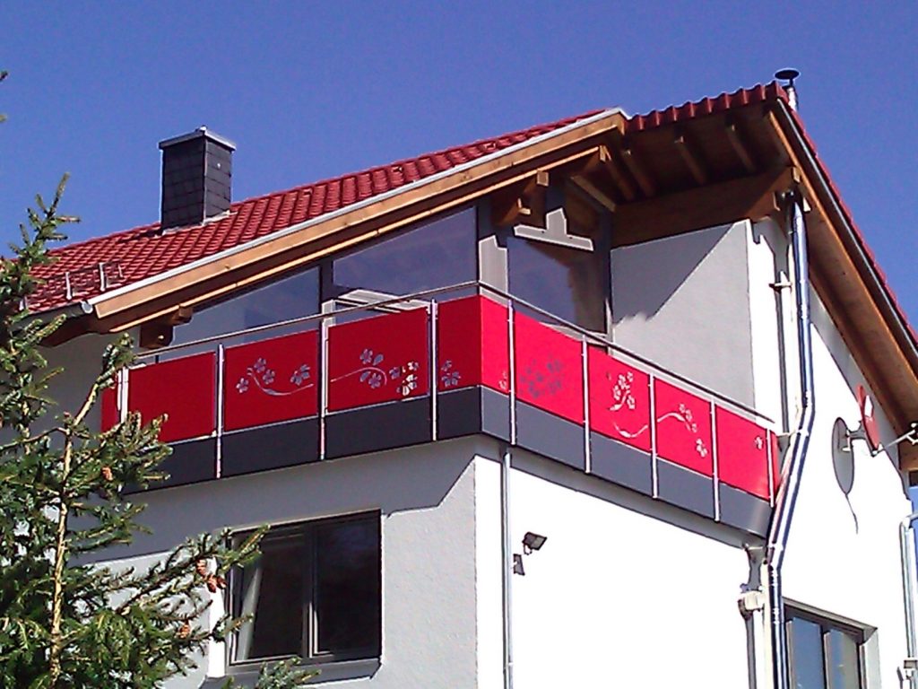 Balkongeländer aus Edelstahlpfosten und Handlauf, mit Füllung aus Aluminiumblechkasetten mit Ausschnitten nach Kundenwunsch und Sockelblende aus Aluminiumblech. Füllbleche rot pulverbeschichtet, Sockelbleche grau pulverbeschichtet